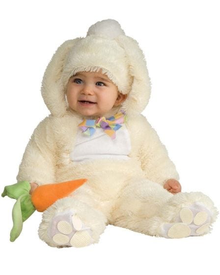 Smiling baby dressed in a fluffy cream-coloured bunny costume with a hood, floppy ears, a pastel bow tie, and holding a plush carrot while sitting on the floor.