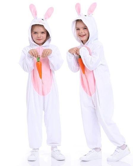 Two young children wearing matching white bunny onesies with pink ears and belly accents, posing playfully, with one holding a plush carrot prop.